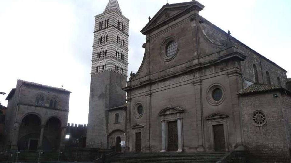 Facciata di una chiesa in pietra con frontone e rosoni circolari, affiancata da un alto campanile quadrato a fasce bianche e scure. Davanti si vedono un’ampia scalinata e una piazza pavimentata; a sinistra è presente un edificio porticato ad archi.