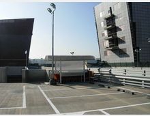 Parcheggio scoperto con stalli delimitati da linee bianche, lampioni alti e una pensilina trasparente al centro. Sullo sfondo si vedono un edificio moderno a più piani con facciata scura e balconi e una lunga struttura arcuata tipo capannone.