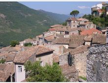 Vista di un borgo collinare con case in pietra addossate tra loro e tetti in coppi di terracotta. Sullo sfondo si vedono pendii montuosi verdi e un albero isolato sopra le abitazioni.