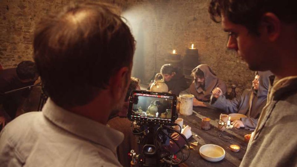Interno in pietra di una taverna o cantina rustica, con volta in muratura e un lungo tavolo di legno illuminato da candele. In primo piano si vede una videocamera con monitor che inquadra la scena del tavolo con stoviglie e brocche.