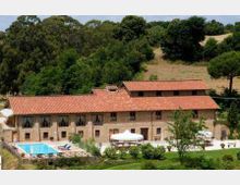 Complesso rurale in pietra con lungo edificio a due piani e tetto in tegole rosse, immerso in un paesaggio collinare con bosco sullo sfondo. In primo piano si vedono una piscina recintata, un’area esterna con ombrelloni bianchi e giardino con alberi e prato.