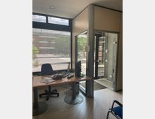 Interno di un piccolo ufficio con scrivania, sedie e monitor, affacciato su grandi finestre con veneziane. Accanto c’è un ingresso vetrato con telaio scuro e una colonna centrale, con vista sulla strada esterna.