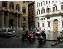 Piccola piazza urbana con pavimentazione in sampietrini bagnati, circondata da palazzi storici in pietra e intonaco con finestre ad arco e portali monumentali. In primo piano sono parcheggiati scooter e un’auto, con dissuasori a sfera lungo il bordo della piazza.