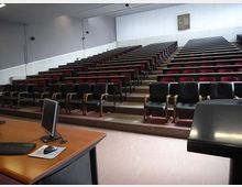 Aula universitaria con gradinate e file di sedili imbottiti rossi e neri disposti a livelli, con pareti chiare e illuminazione a soffitto. In primo piano si vede un banco docente con computer e, sul lato, un leggio nero.