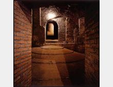 Corridoio sotterraneo in mattoni e pietra con soffitto a volta e una serie di archi, illuminato da una lampada a parete. Pavimento in pietra con gradini circolari e passaggio che conduce a un’apertura ad arco in fondo.