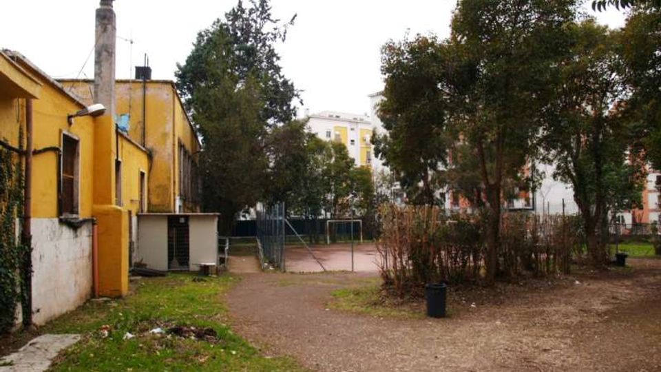 Cortile urbano con area sterrata e una rete metallica che delimita un piccolo campo sportivo; a sinistra si vede un edificio basso giallo con camino. Alberi e cespugli circondano lo spazio, con palazzi residenziali sullo sfondo.