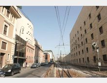 Strada urbana rettilinea con binari ferroviari e cavi aerei sul lato destro, affiancata da un lungo edificio in mattoni con piccole finestre. Sul lato sinistro si vedono palazzi moderni e storici, auto parcheggiate e lampioni lungo il marciapiede.