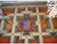Soffitto decorato di un edificio storico con motivi geometrici a riquadri in tonalità rosso e oro, cornici ornate e dettagli a rosette. Al centro è presente uno stemma araldico in rilievo, con lampadari bianchi ai lati.