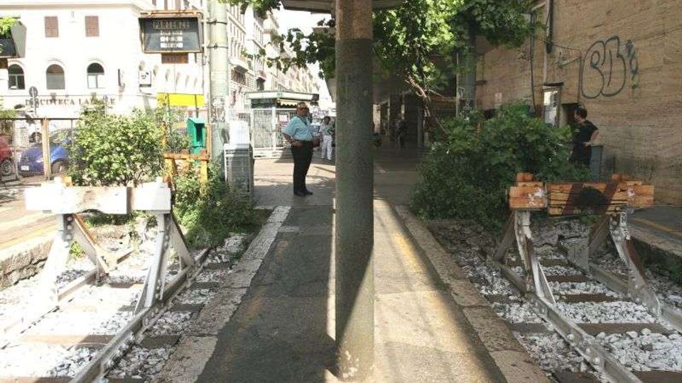 Pensilina di una fermata del tram lungo un marciapiede stretto, con binari dismessi ai lati e traversine tra ghiaia bianca. Sullo sfondo si vedono edifici urbani, vegetazione spontanea vicino ai binari e un muro con graffiti.