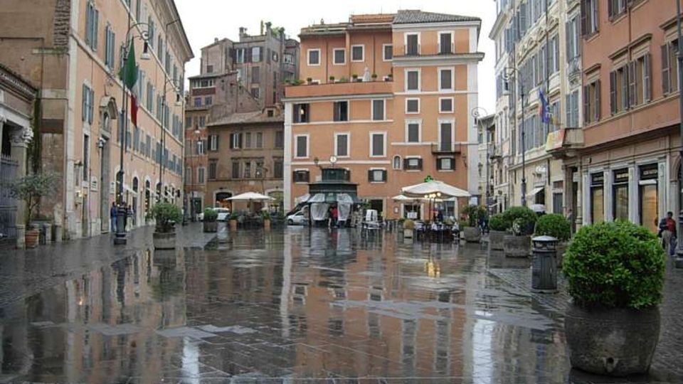 Piazza urbana con pavimentazione in pietra bagnata dalla pioggia, che riflette gli edifici circostanti. Intorno si affacciano palazzi storici dai toni ocra e pastello con persiane e negozi al piano terra, con tavolini sotto ombrelloni e grandi fioriere lungo il perimetro.