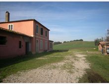 Cascina di campagna con edificio intonacato rosa a due piani, tetto in coppi e persiane scure, affacciata su un cortile in ghiaia e prato. Sullo sfondo si vedono campi coltivati ondulati e un piccolo annesso rustico ai margini della proprietà.