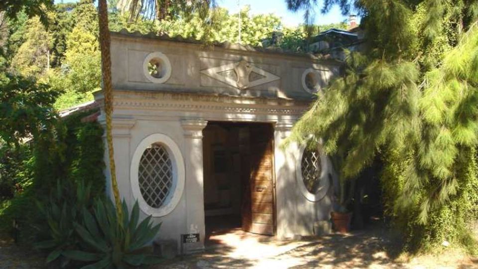 Piccolo edificio in muratura in un giardino, con facciata classica decorata, ingresso centrale con porta in legno e due finestre ovali con griglie. Intorno ci sono alberi alti, palme e vegetazione fitta che ombreggia il vialetto d’accesso.