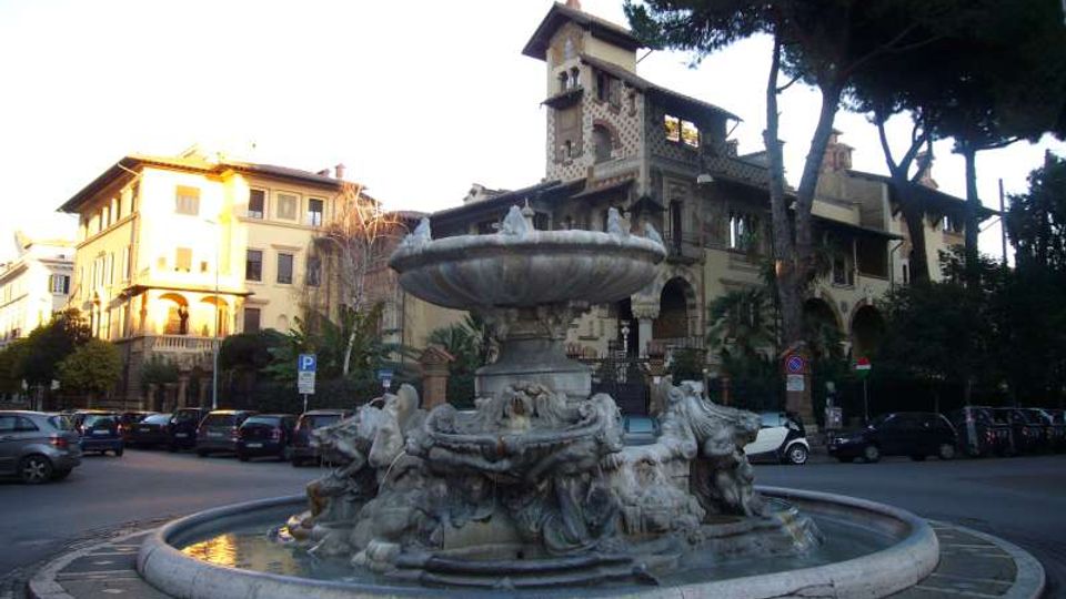 Fontana monumentale in una rotonda, con grande vasca circolare e sculture in pietra attorno al basamento. Sullo sfondo si vedono edifici storici con torretta e portici, alberi alti e auto parcheggiate lungo la strada.