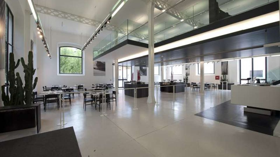 Ampio interno in stile industriale con pavimento chiaro, soffitto alto e soppalco con balaustra in vetro sostenuto da colonne. Lungo le pareti ci sono grandi finestre, illuminazione a binario e tavoli con sedie disposti come area ristorante o lounge.