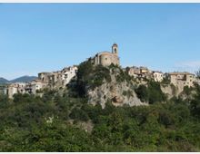 Veduta panoramica di un borgo storico arroccato su uno sperone roccioso, con una chiesa in pietra e campanile in cima e case addossate lungo i fianchi. In primo piano si estende una vegetazione verde, con colline lontane sotto un cielo sereno.