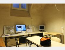 Piccola aula o sala studio con pareti in intonaco e soffitto a volta, illuminata da una finestra alta. Lungo un tavolo sono presenti postazioni con computer e materiali informativi, con banchi e sedie disposti davanti.