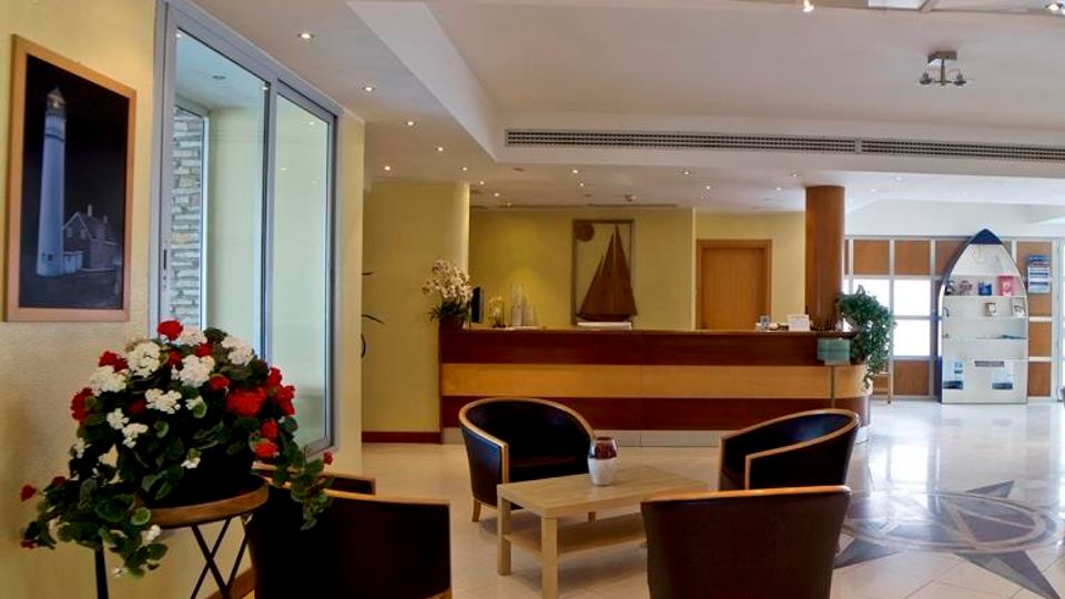 Hall di un hotel con pavimento in piastrelle lucide e area salotto con quattro poltrone scure e tavolino basso al centro. Sullo sfondo si vede il banco reception in legno, illuminazione a faretti nel controsoffitto, vetrate laterali e decorazioni come un grande vaso di fiori e quadri alle pareti.