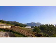 Paesaggio collinare rurale con campi coltivati in pendenza, recinzioni e alcuni edifici agricoli sulla sinistra. Sullo sfondo si vedono montagne e una valle con banchi di nebbia tra alberi e bosco sotto un cielo limpido.