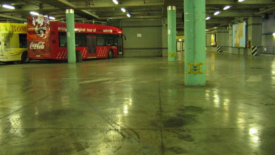 Ampio garage sotterraneo con pavimento in cemento lucido e segni di pneumatici, illuminato da file di lampade al neon e sostenuto da colonne verdi. Sul lato sinistro è parcheggiato un autobus rosso accanto a pareti grigie e cartelloni pubblicitari.