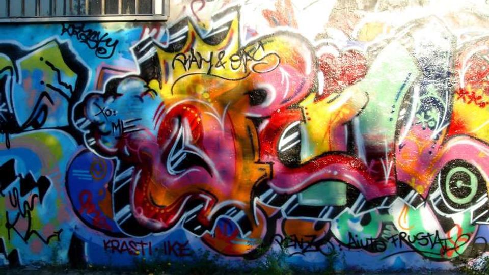 Muro esterno di un edificio ricoperto da un grande murale di graffiti multicolori con forme astratte e scritte sovrapposte. In alto è visibile una finestra con inferriate, mentre alla base del muro ci sono erba e vegetazione.
