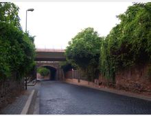 Strada in pavé che passa sotto un ponte e un viadotto, con un sottopasso ad arco in muratura sullo sfondo. Ai lati ci sono muri in mattoni e scarpate ricoperte di vegetazione, con marciapiede e un lampione.