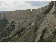 Ripida parete di falesia rocciosa e friabile, con creste erose e canaloni verticali, punteggiata da piccoli cespugli. Sullo sfondo si vedono altre formazioni collinari sotto un cielo nuvoloso.