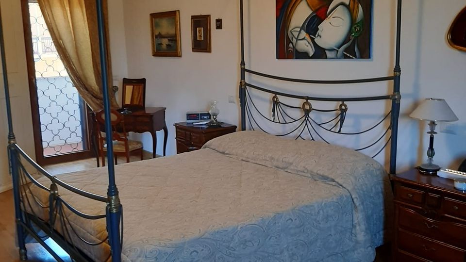 Camera da letto con letto matrimoniale a baldacchino in ferro battuto e copriletto chiaro, affiancato da comodini in legno con lampada. Sulla parete è appeso un grande quadro colorato e a sinistra c’è una portafinestra con tende che dà su un balcone con ringhiera.