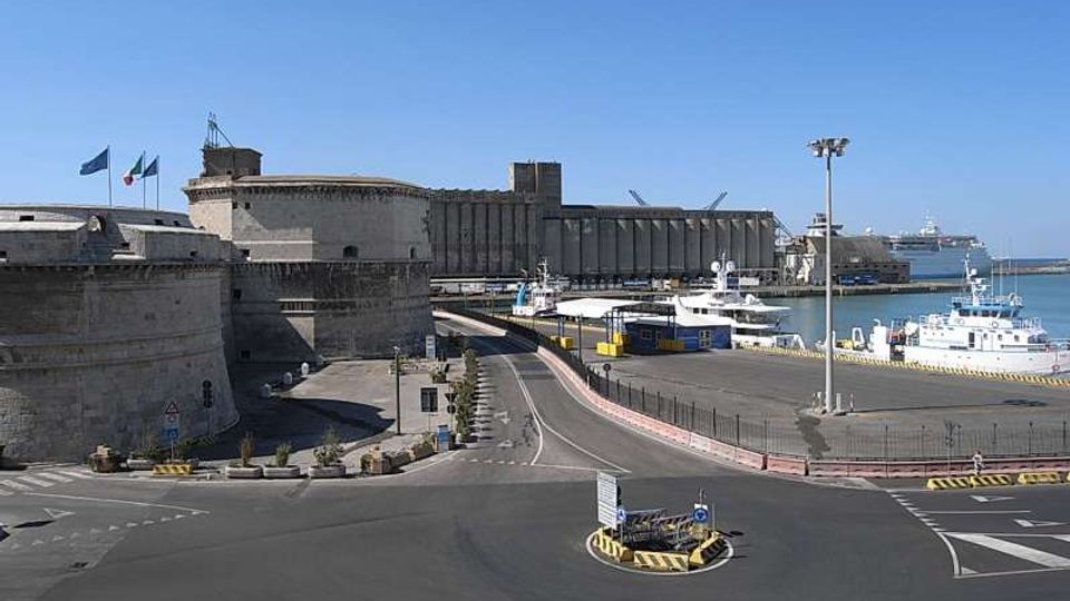 Veduta di un’area portuale con banchine e traghetti attraccati, affiancata da una grande fortificazione in pietra con mura e torri. In primo piano si vede una strada con rotatoria e segnaletica, con il mare e l’orizzonte sullo sfondo.