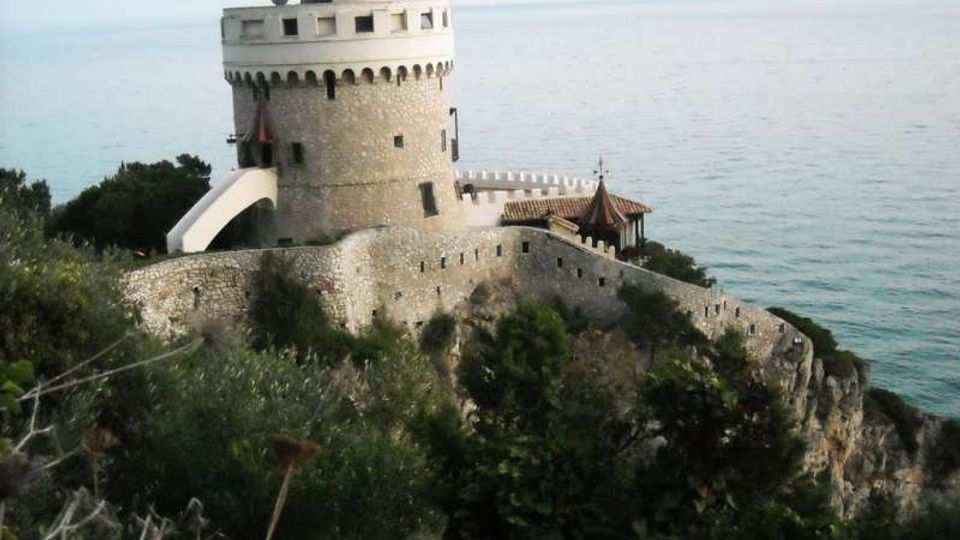 Torre fortificata in pietra con corona merlata e piccola cupola bianca, collegata a mura difensive su un promontorio roccioso. Sullo sfondo si vede il mare aperto, con vegetazione mediterranea in primo piano.