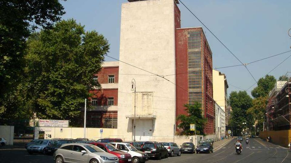 Edificio industriale alto con grande torre bianca e struttura laterale in mattoni rossi con finestre verticali, affacciato su una strada urbana. In primo piano ci sono un parcheggio con molte auto e binari del tram con cavi aerei, circondati da alberi.