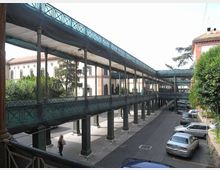 Passerella sopraelevata in metallo con struttura reticolare e tettoia, sostenuta da una lunga fila di pilastri, che corre lungo una strada con auto parcheggiate. Sullo sfondo si vedono edifici in muratura e aree alberate in un contesto urbano.