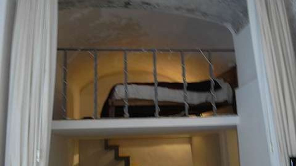 Interno di un piccolo appartamento con soppalco: in basso zona soggiorno con tavolo rotondo e scala a gradini che porta al livello superiore. Il soppalco ha una ringhiera metallica e un soffitto ad arco in pietra, con tende chiare ai lati dell’apertura.