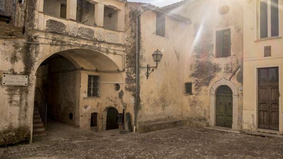 Piccola piazza lastricata in pietra circondata da edifici storici in intonaco consumato, con un passaggio ad arco e loggiato superiore, lanterna a muro e portali in legno. La luce del sole entra dall’angolo in alto a destra, evidenziando le facciate con finestre e persiane.