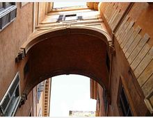 Vista dal basso di un passaggio ad arco che collega due edifici storici in muratura color ocra, con finestre e persiane sui lati. L’arco crea un corridoio coperto sopra la strada e incornicia un tratto di cielo luminoso.