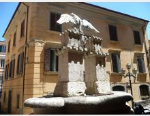 Fontana in pietra al centro di una piccola piazza, con basamento circolare e due pilastri sormontati da una scultura di aquila con le ali spiegate. Sullo sfondo si vede un edificio storico color ocra con persiane scure e lampioni in ferro.