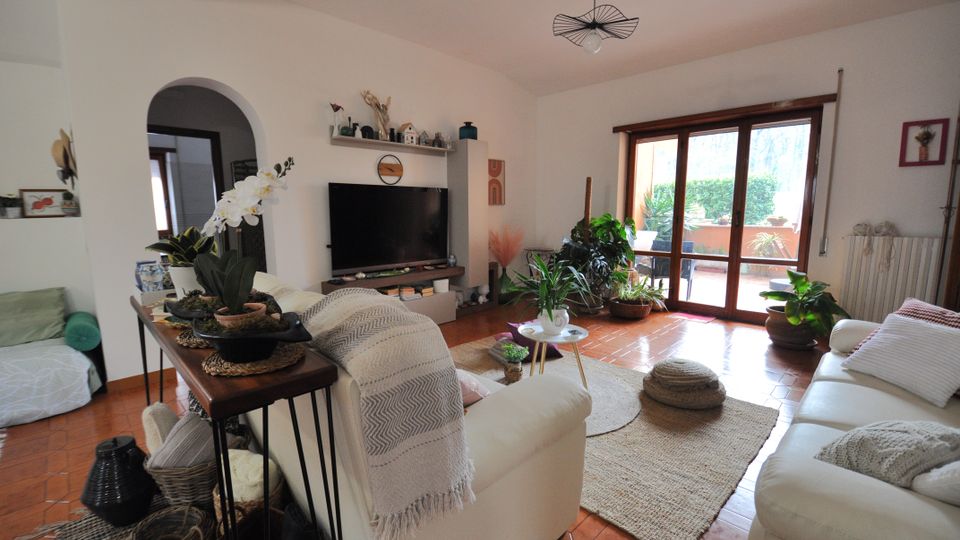 Soggiorno luminoso con pavimento in cotto, divani chiari e tappeto centrale, con mobile TV e arco che conduce a un’altra stanza. Ampia portafinestra in legno che si apre su un terrazzo con piante, con diverse piante da interno vicino alla finestra.