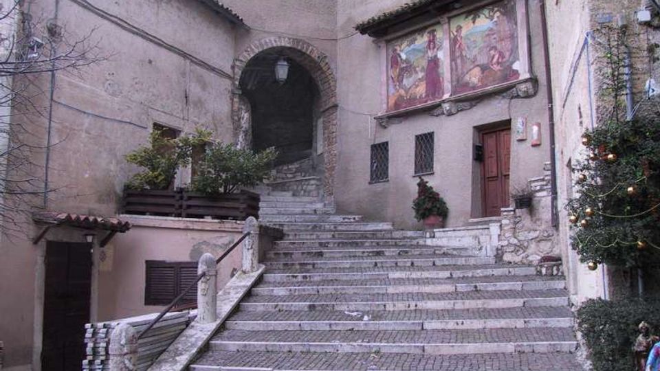 Scalinata in pietra e mattoni che sale in un cortile storico, con archi in muratura e facciate intonacate. Sulla parete a destra sono visibili un grande affresco, una porta in legno e piccole finestre con inferriate, con piante e decorazioni lungo i lati.