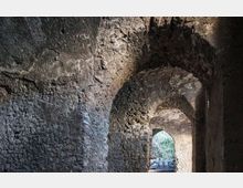 Interno di un corridoio in pietra grezza con volte ad arco e pareti irregolari. In fondo si apre un arco che conduce verso l’esterno, dove si intravede vegetazione e luce naturale.