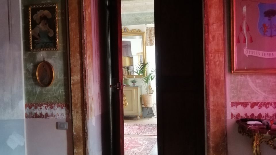 Interno di una casa storica con pareti decorate e cornice di porta in legno dipinto; una doppia porta socchiusa conduce a una stanza luminosa con tappeti e mobili. Sulle pareti sono appesi quadri e fotografie, e a destra si vede una piccola consolle con dettagli ornamentali.