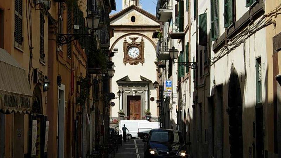 Strada stretta di centro storico fiancheggiata da edifici alti con persiane verdi, balconi e lampioni a muro. In fondo alla via si vede la facciata di una chiesa con orologio centrale e campanile, con alcune auto parcheggiate lungo la carreggiata.