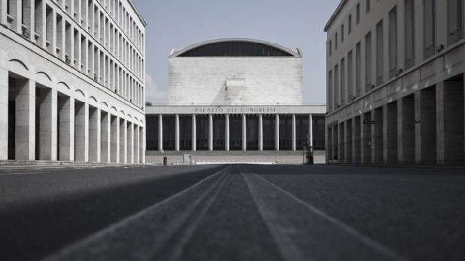 Ampia piazza lastricata tra due edifici simmetrici con portici ad arcate, che incorniciano un grande edificio monumentale sullo sfondo con facciata in pietra, alte colonne e tetto curvo. Vista centrale prospettica lungo l’asse della piazza, con lampioni e spazio aperto privo di arredi.