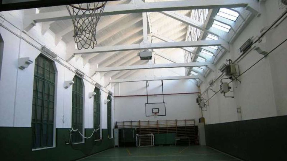 Interno di una palestra scolastica con campo da basket, pareti bianche e verdi e grandi finestre ad arco su un lato. Il soffitto alto con travi a vista e lucernari lungo la falda illumina lo spazio; si vedono canestri e attrezzature sportive sul fondo.