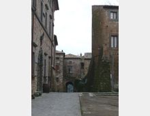 Strada acciottolata in un borgo storico, fiancheggiata da alti edifici in pietra con finestre e tetti in coppi. In fondo si vede un arco in muratura che attraversa il passaggio, con muri segnati da muschio e umidità.