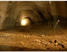 Interno di un tunnel sotterraneo con pareti e soffitto in terra compatta, illuminato da una luce intensa visibile in fondo. Il pavimento è polveroso e irregolare, con piccoli sassi e rami secchi in primo piano.