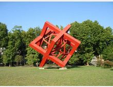 Grande scultura astratta in metallo rosso, composta da telai cubici inclinati e intrecciati, collocata su un prato. Sullo sfondo si vede un’area verde con alberi ad alto fusto e cielo sereno.