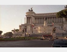Grande monumento neoclassico in marmo bianco con lunga facciata a colonne, ampie scalinate e statue equestri in cima, affacciato su una rotonda con traffico. Davanti si vedono aiuole e bandiere, con alberi ai lati e cielo al tramonto sullo sfondo.