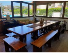 Sala da pranzo luminosa con grande tavolo in legno e panche, affiancata da divano e tastiera elettronica, circondata da ampie finestre angolari. Dalle vetrate si vede un giardino con siepi e una piscina all’esterno.