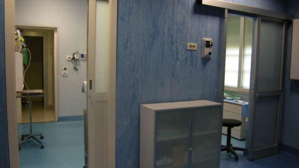 Interno di una struttura sanitaria con pavimento blu e pareti azzurre, con porte scorrevoli e un armadietto metallico. Sullo sfondo si vedono un corridoio con attrezzature mediche e una piccola stanza con finestra e arredi da lavoro.