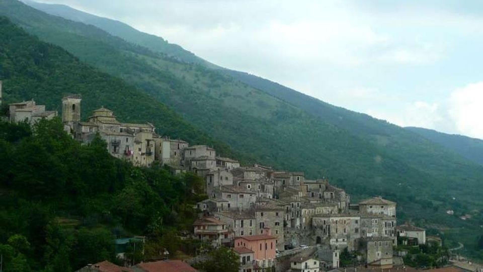Borgo di montagna con case in pietra addossate al pendio, dominato da una chiesa e un campanile. Sullo sfondo si vedono colline boscose e montagne sotto un cielo nuvoloso.