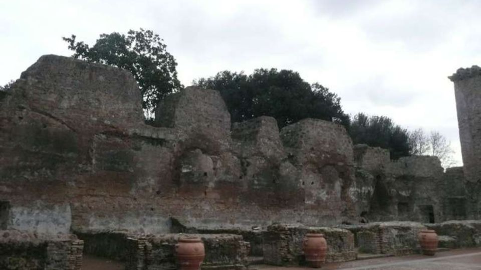 Resti di un antico muro in pietra e mattoni, con ampie porzioni crollate e superfici erose, in un’area archeologica all’aperto. In primo piano si vede una piazza lastricata con muretti bassi e grandi anfore, con alberi sullo sfondo sotto un cielo nuvoloso.
